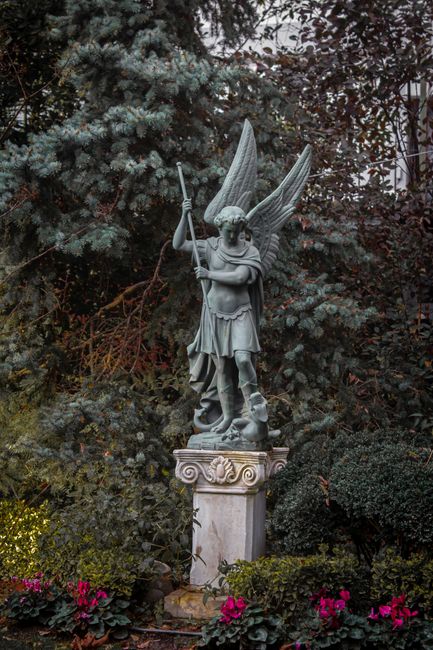 A stone statue of an angel with large, detailed wings, standing on a decorated pedestal in a garden setting. The angel is holding a staff in its right hand and has a serene expression. Surrounding the
