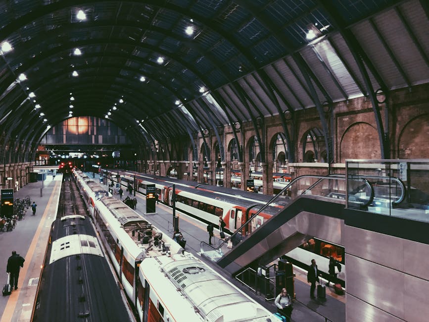 The image depicts a large indoor train station with a high, arched metal roof supported by brick and iron columns. Multiple train tracks run parallel, with several modern passenger trains stationed al