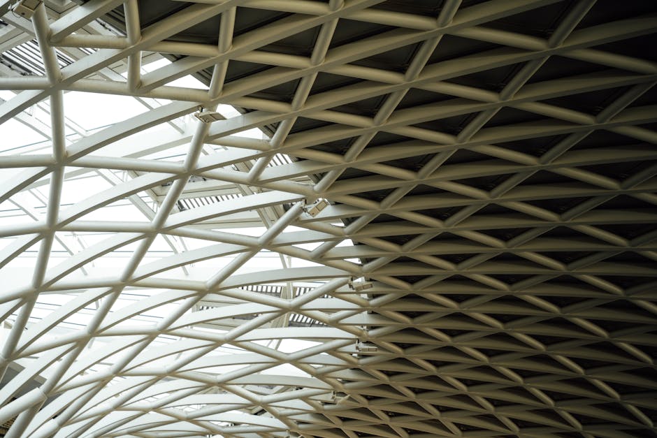 The image depicts a large indoor train station with a high, arched metal roof supported by brick and iron columns. Multiple train tracks run parallel, with several modern passenger trains stationed al
