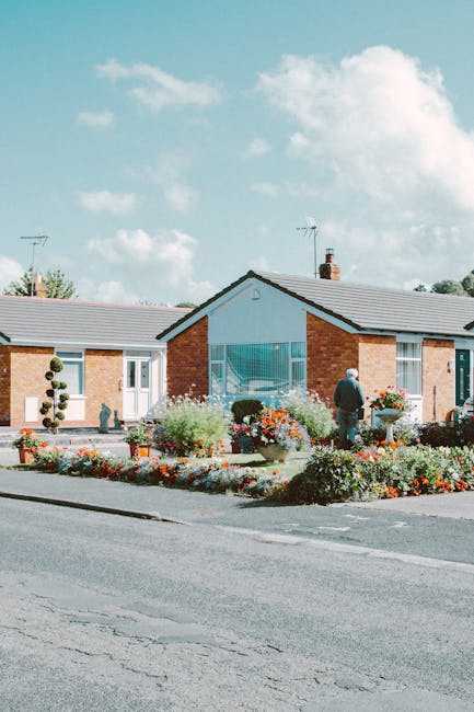 A single-story residential building with a pitched roof and brick exterior walls, situated along a paved street. In front of the house, there is a well-maintained garden bed filled with colorful flowe